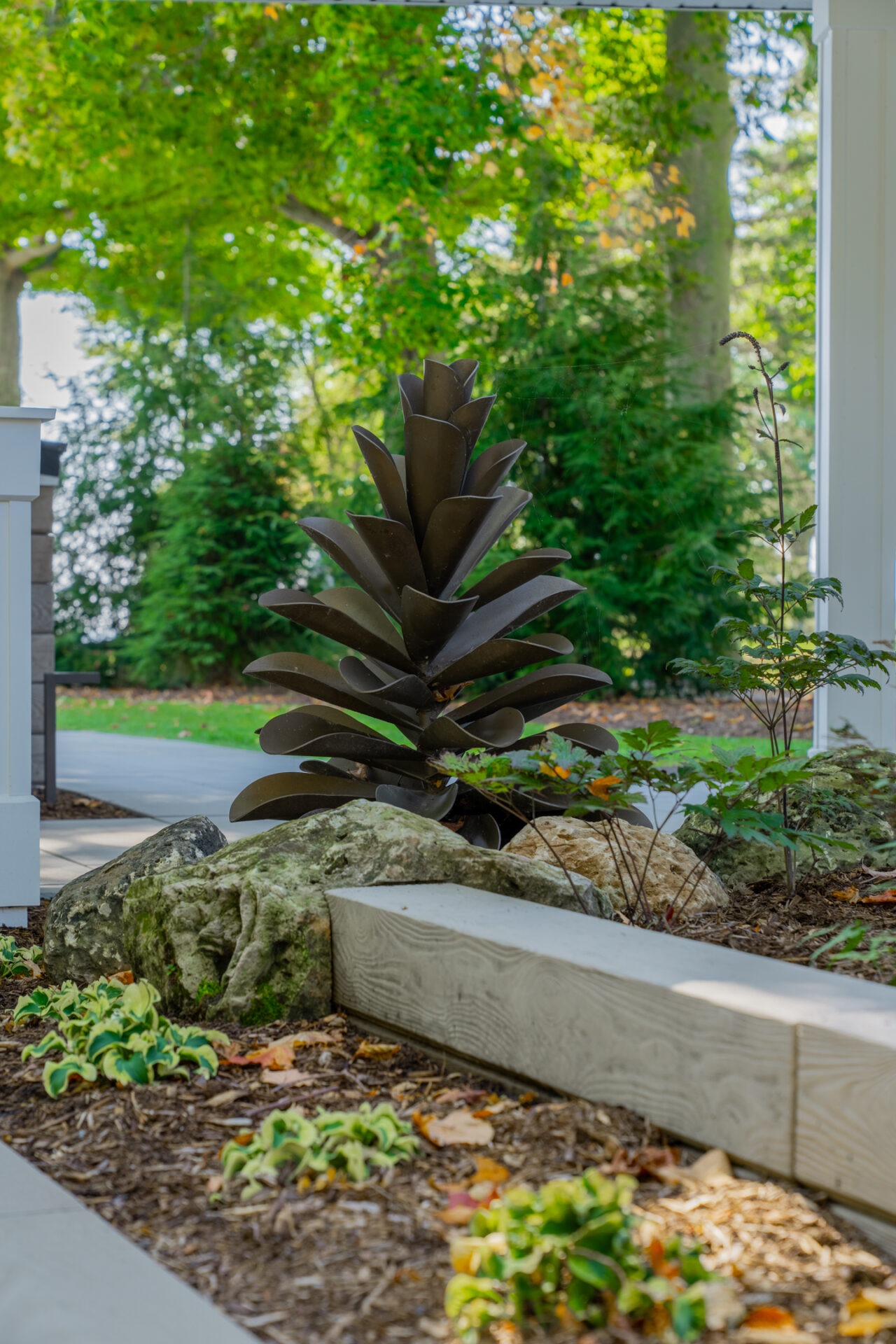 Close-up of a decorative metal pinecone sculpture in a landscaped garden bed with mulch and rocks.