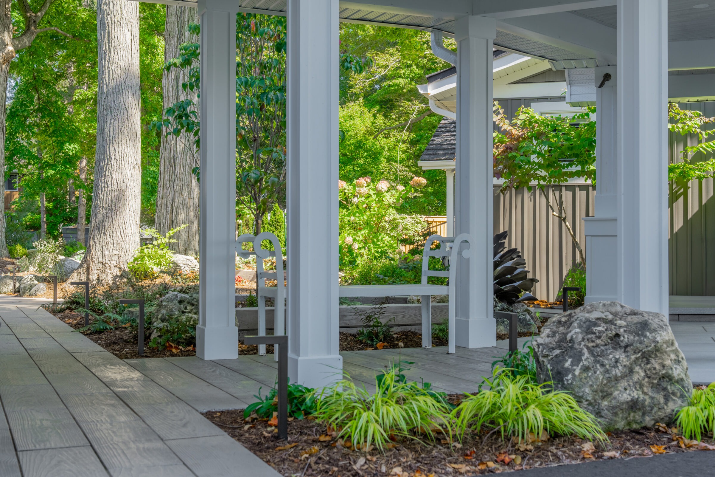A view from under a white covered walkway featuring stone paths, garden beds, and a white bench.