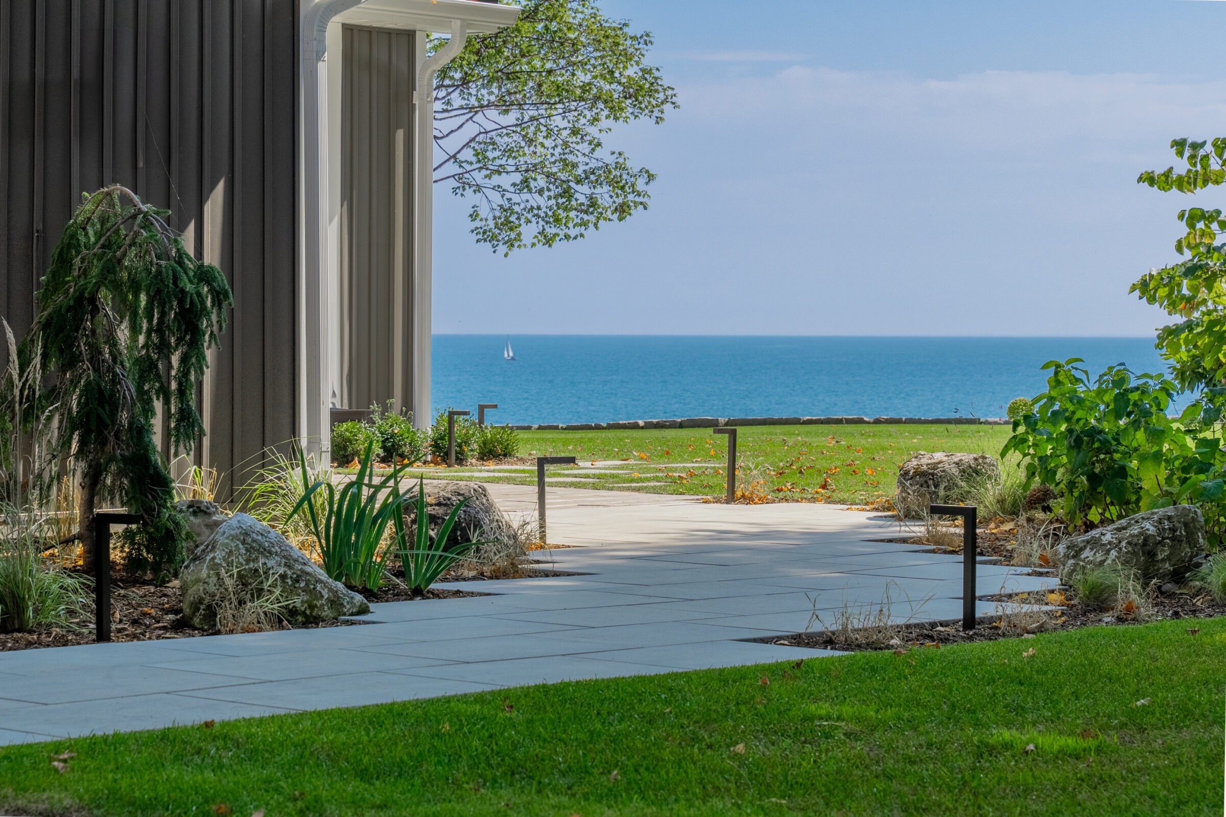 A clean stone walkway leading toward a vast blue lake with a small sailboat visible on the horizon.