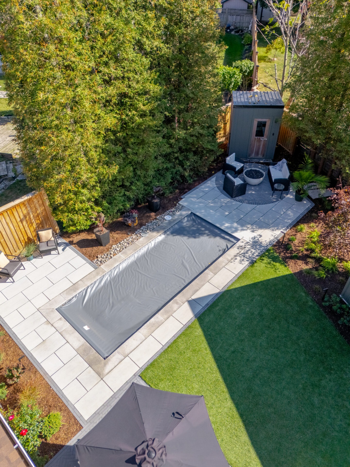 High-angle view of a backyard featuring a rectangular pool with a grey cover, stone patio, and modern sauna.