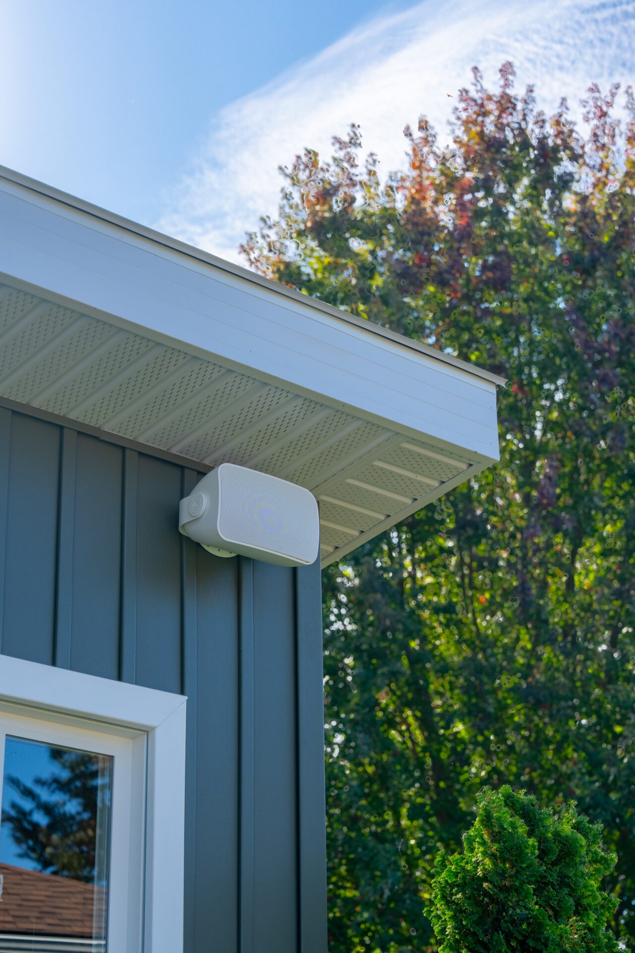 White outdoor speaker mounted to the dark green soffit of a pool house.