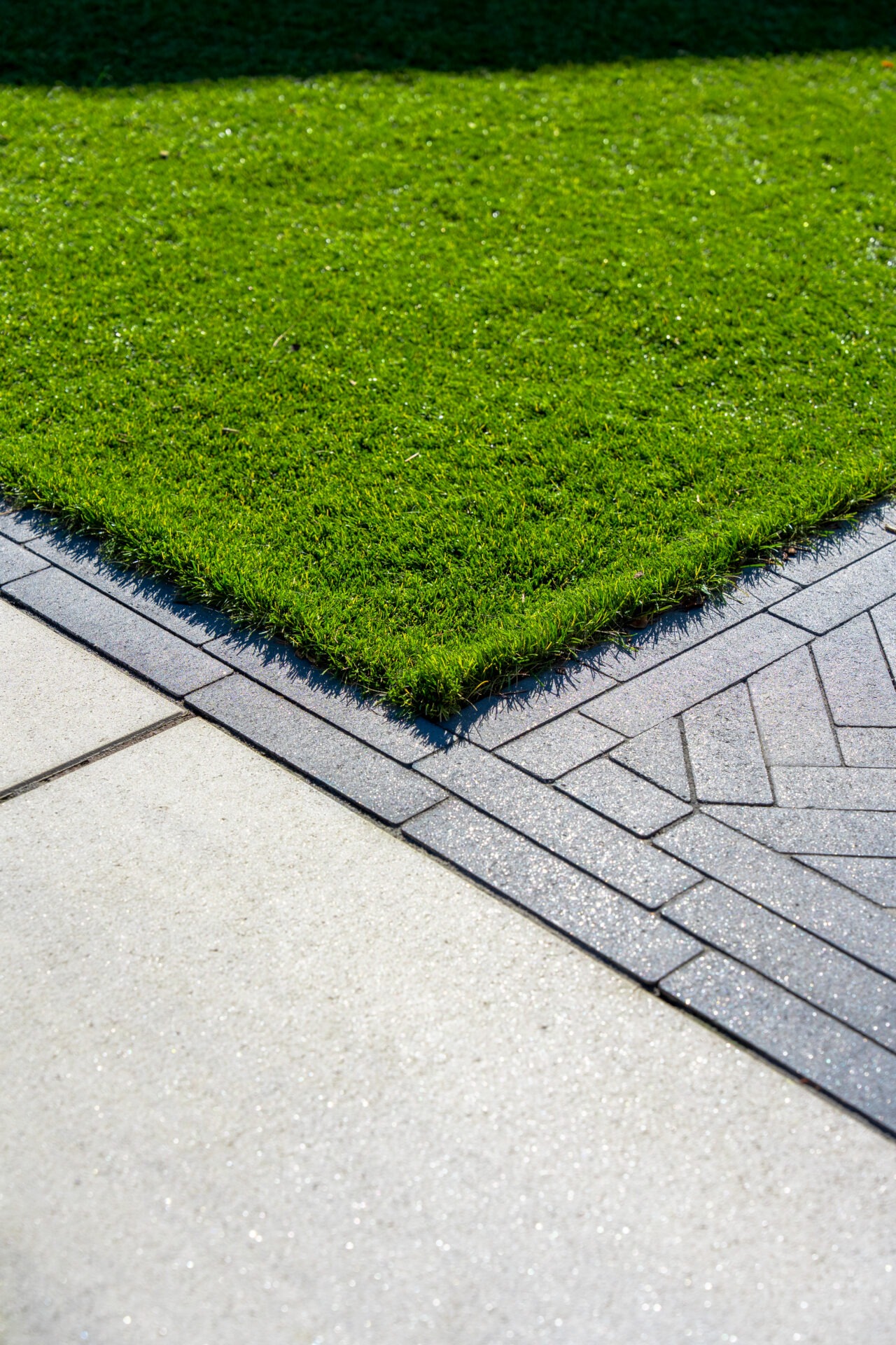 A top-down detail shot of a sharp corner where a vibrant green lawn meets a dark grey brick border. The border transitions into a light grey stone patio on one side and a dark grey herringbone-patterned walkway on the other.