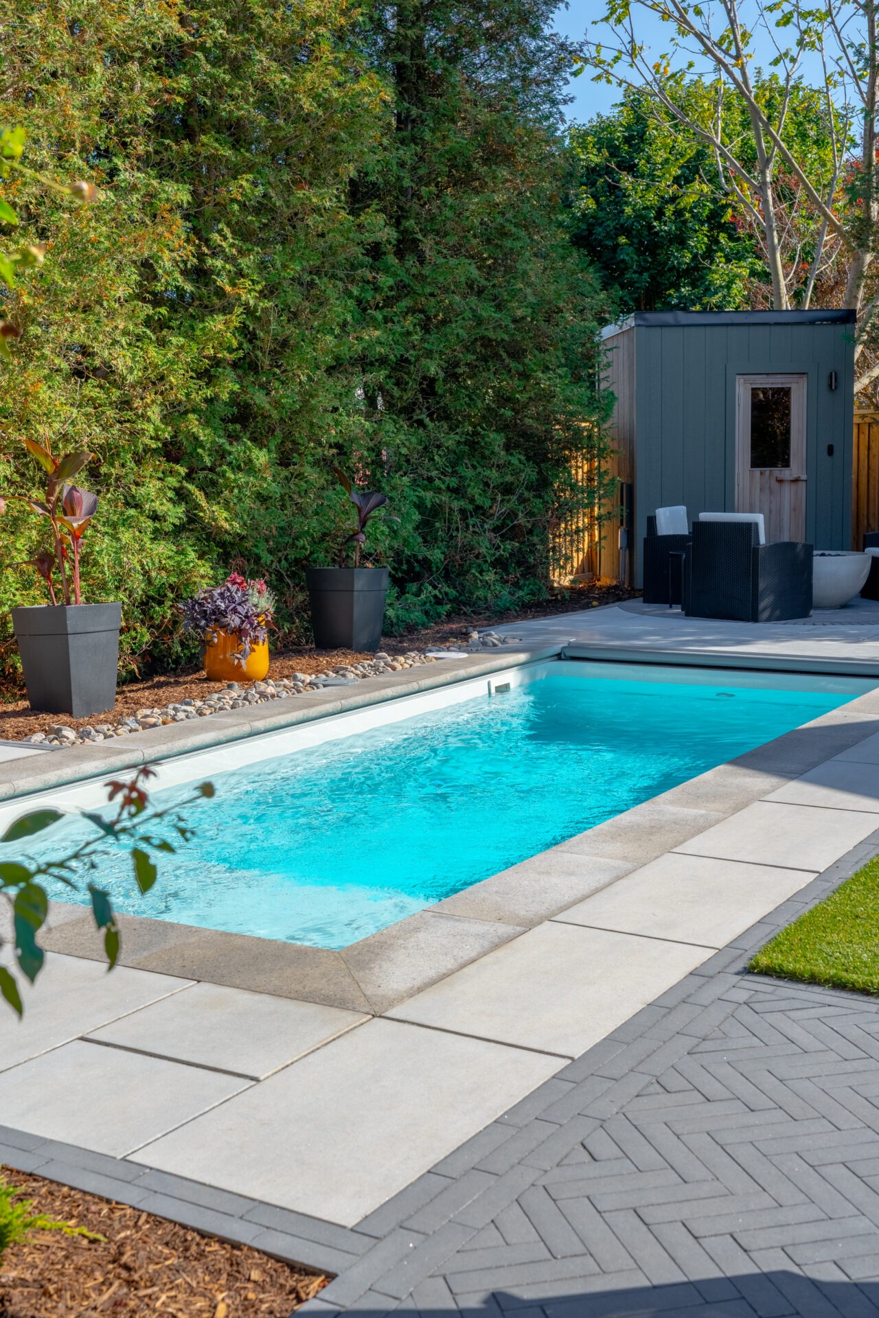 Daytime view of a rectangular pool with stone coping, black planters, and a green sauna building.