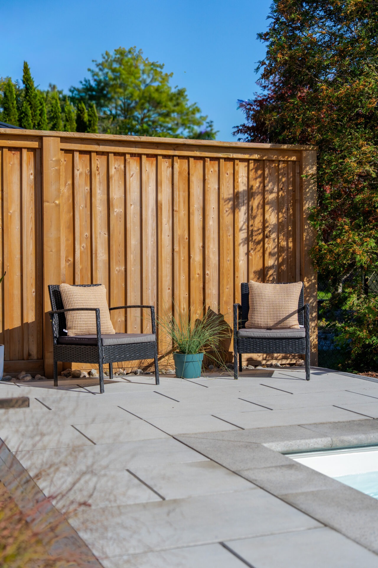 Two black wicker chairs with beige pillows on a stone patio against a vertical wood privacy fence.