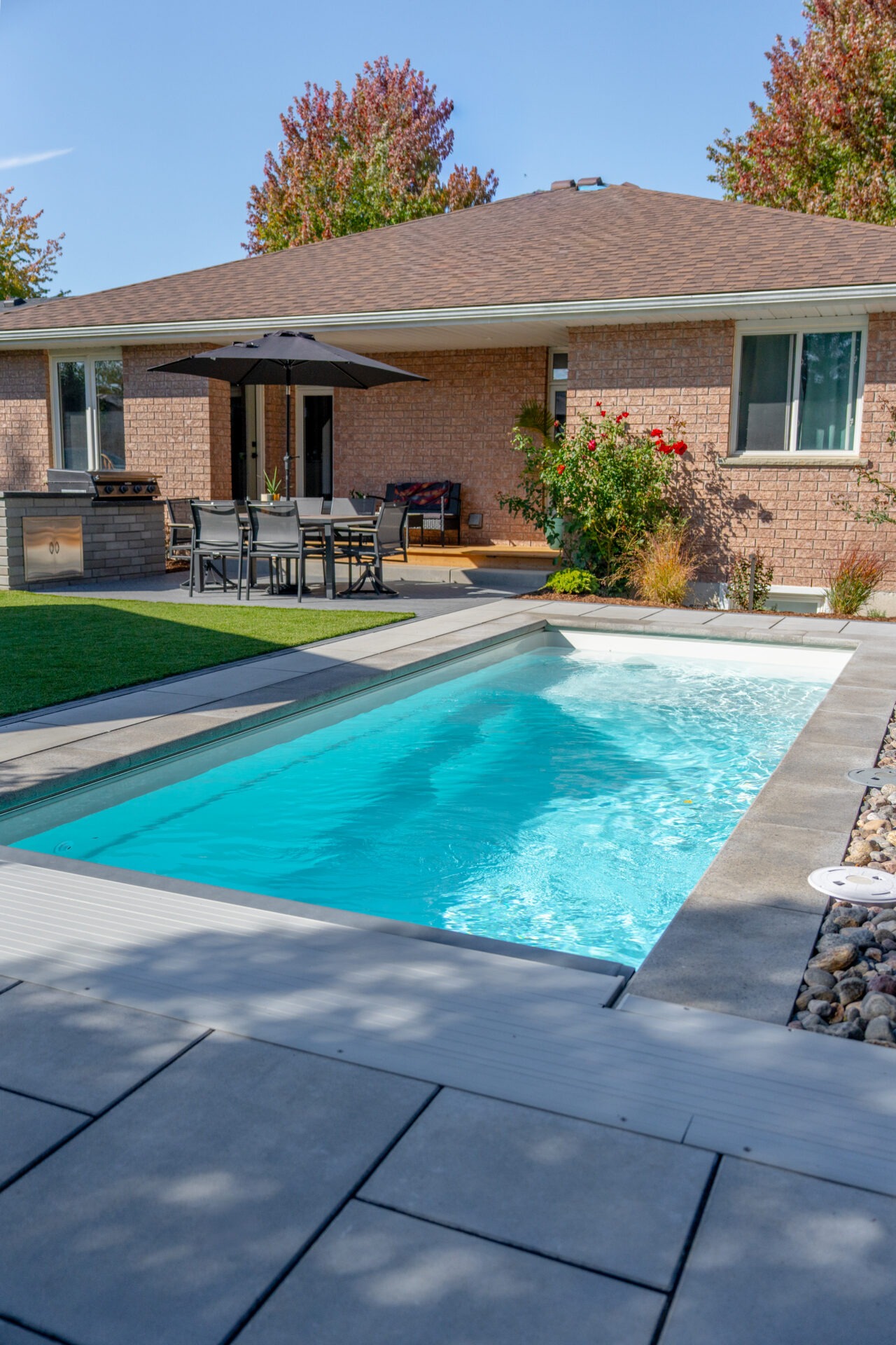 Backyard view of a bright blue rectangular pool, stone patio, and a brick house with an outdoor dining area.