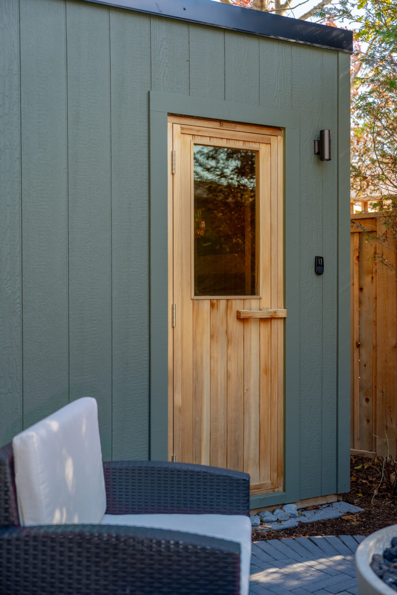Close-up of a modern green sauna with a natural wood door and glass window, featuring sleek black exterior lighting.