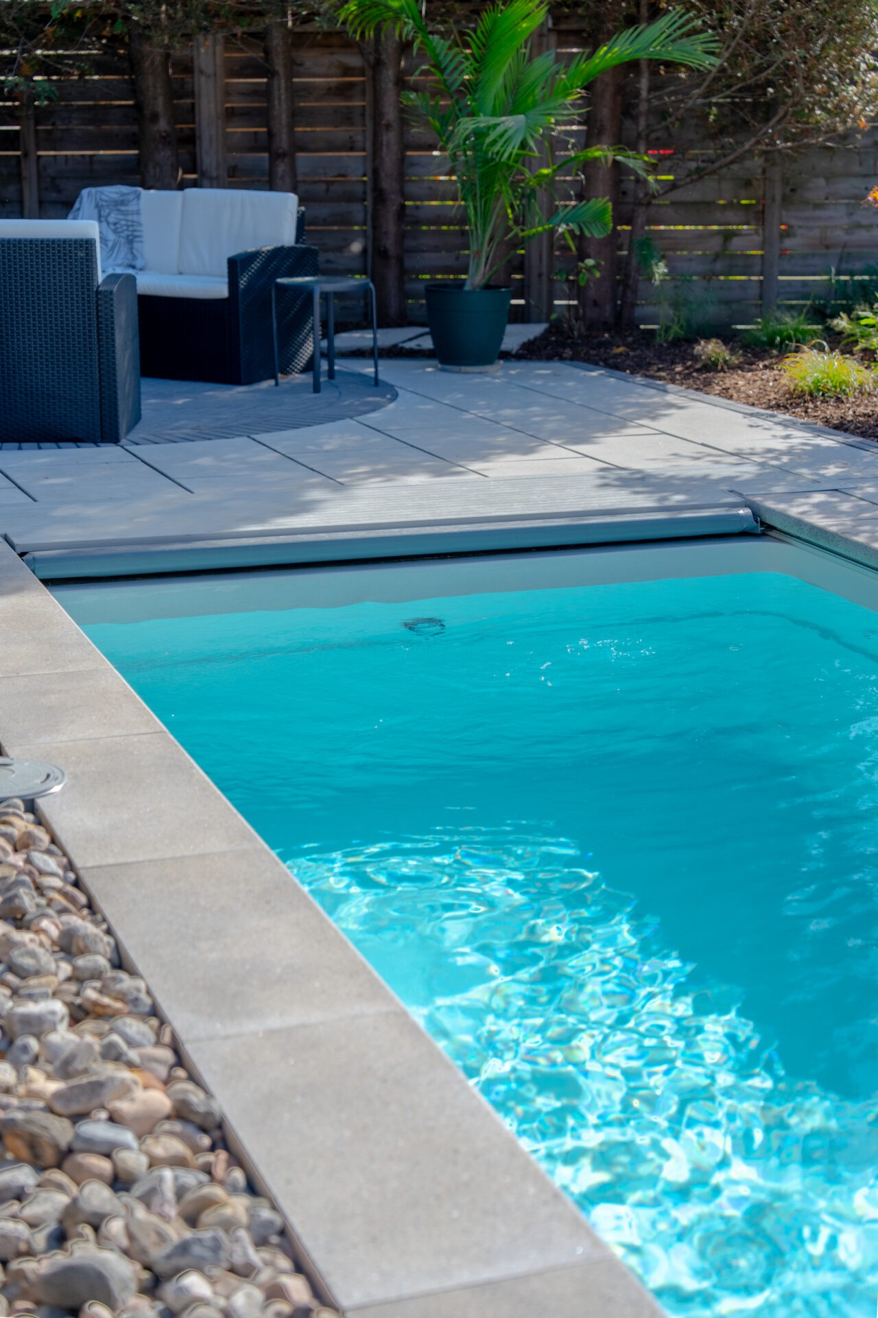 View of a bright turquoise inground pool with stone coping next to a lounge area with black wicker furniture.