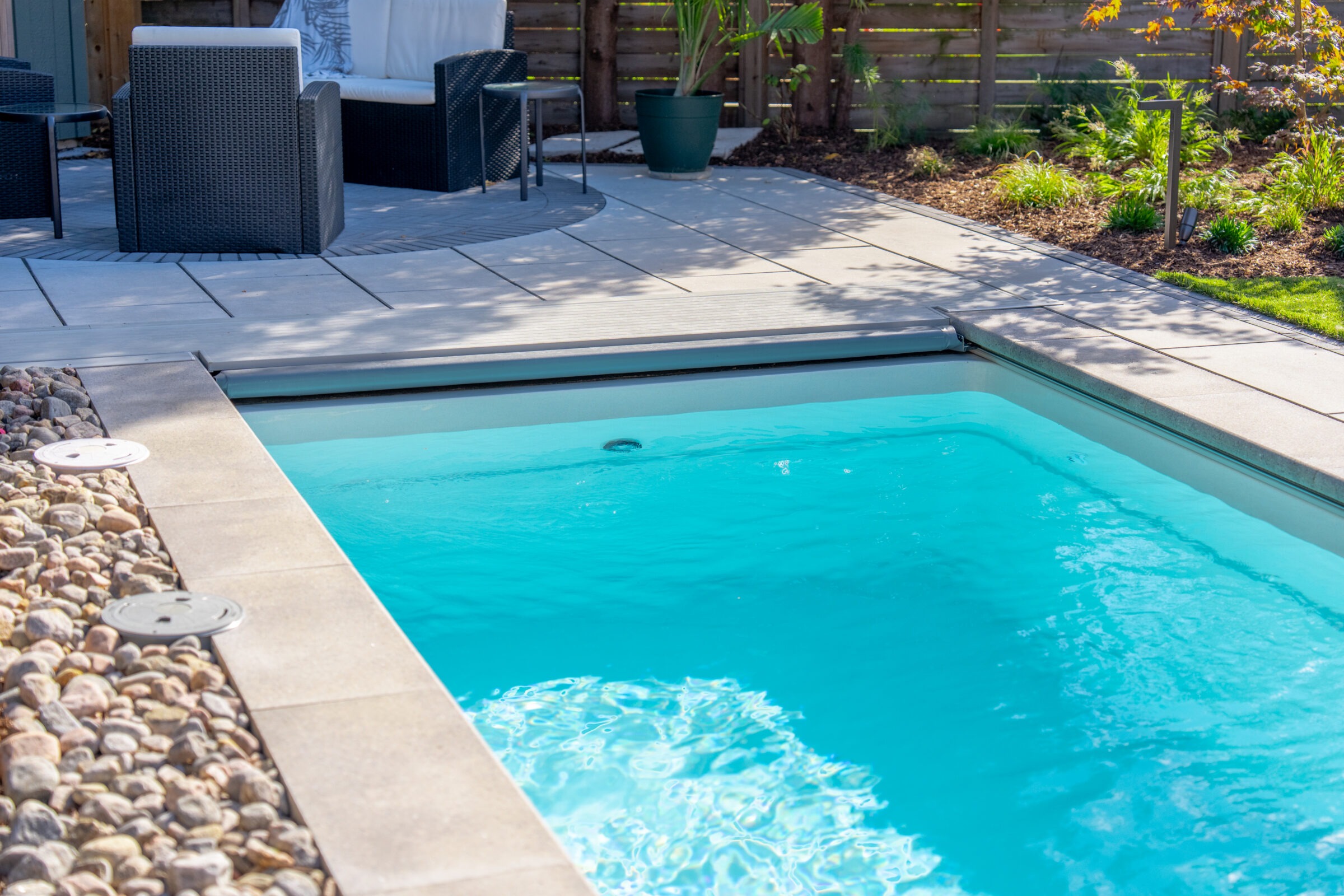 Ground-level view of an inground pool with a retracted grey cover, surrounded by stone pavers and garden mulch.