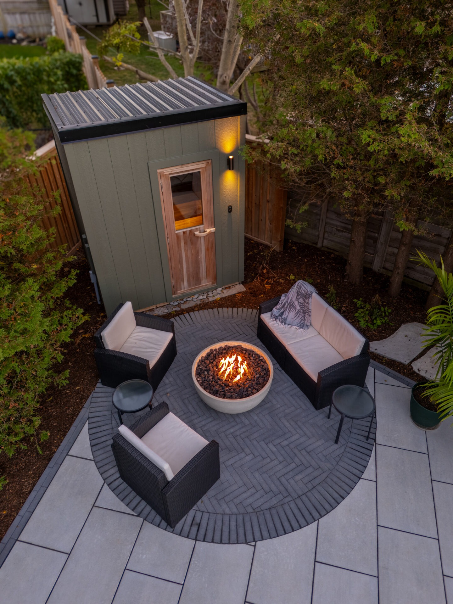 Overhead view of a circular fire pit lounge with black wicker furniture and a modern green sauna building.