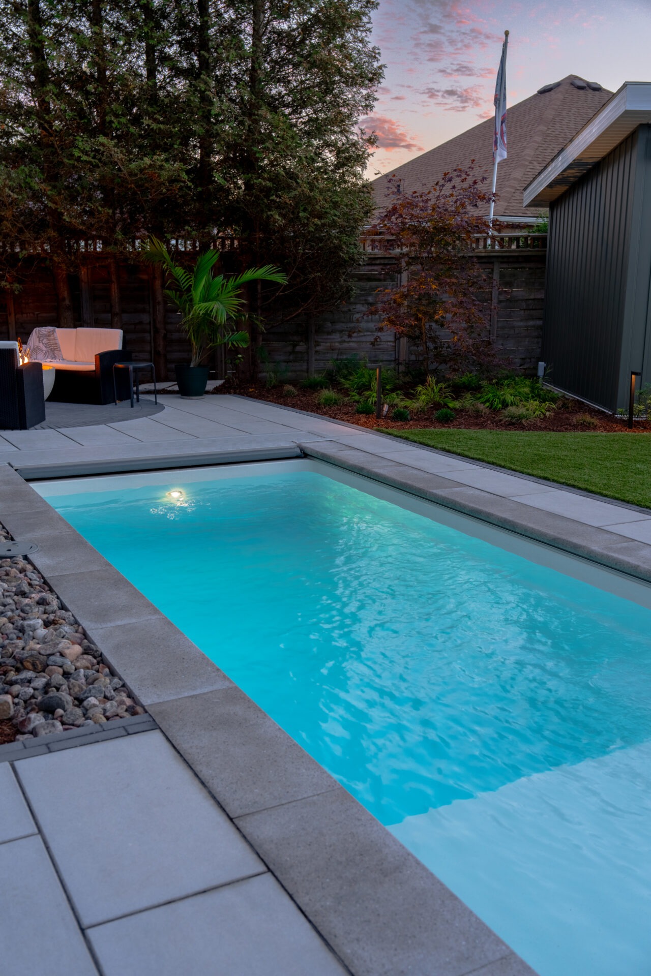 Night view of a glowing turquoise inground pool and stone patio under a pink sunset sky.