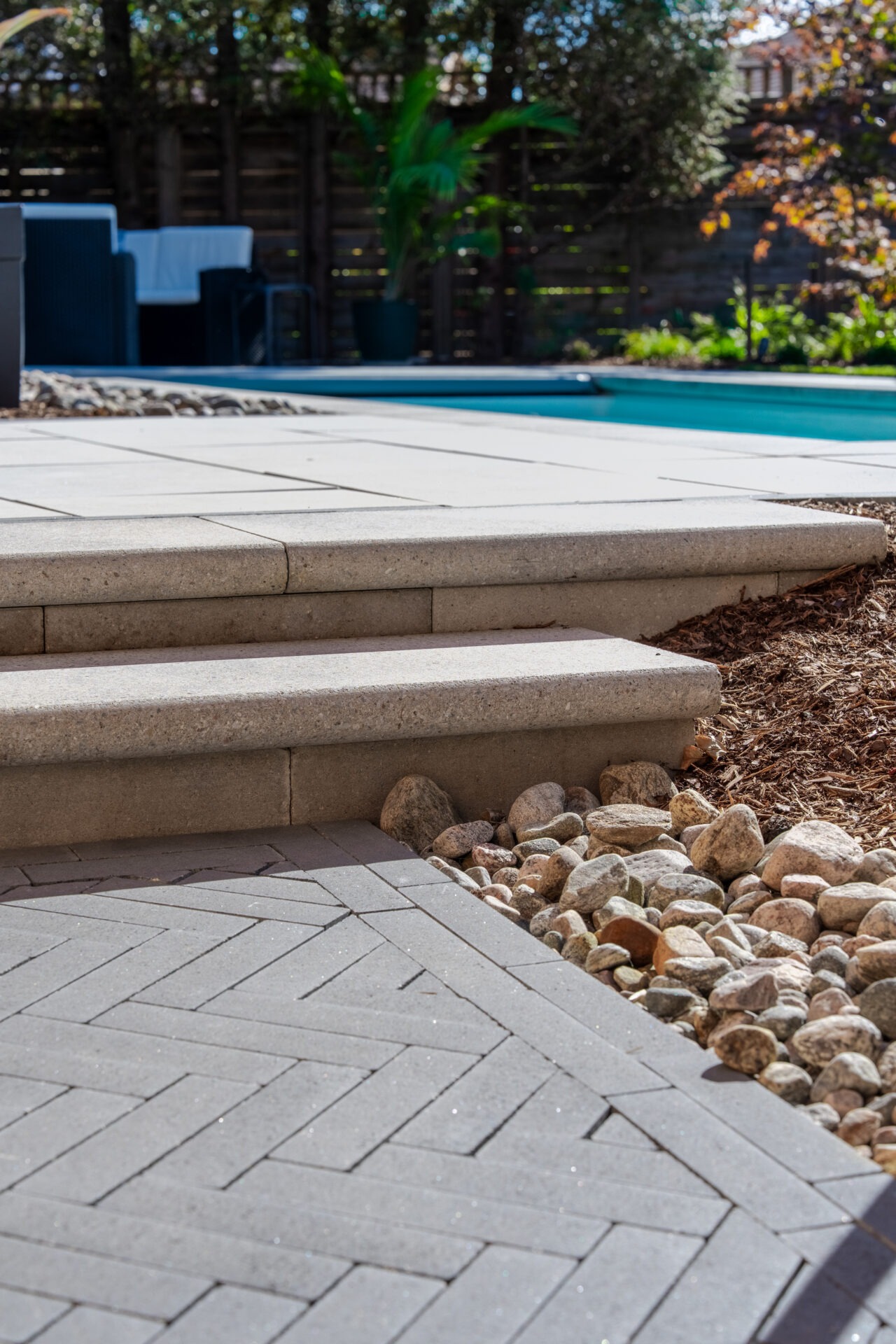 Close-up of light grey stone steps transitioning into a dark grey herringbone paver walkway with river rock accents.