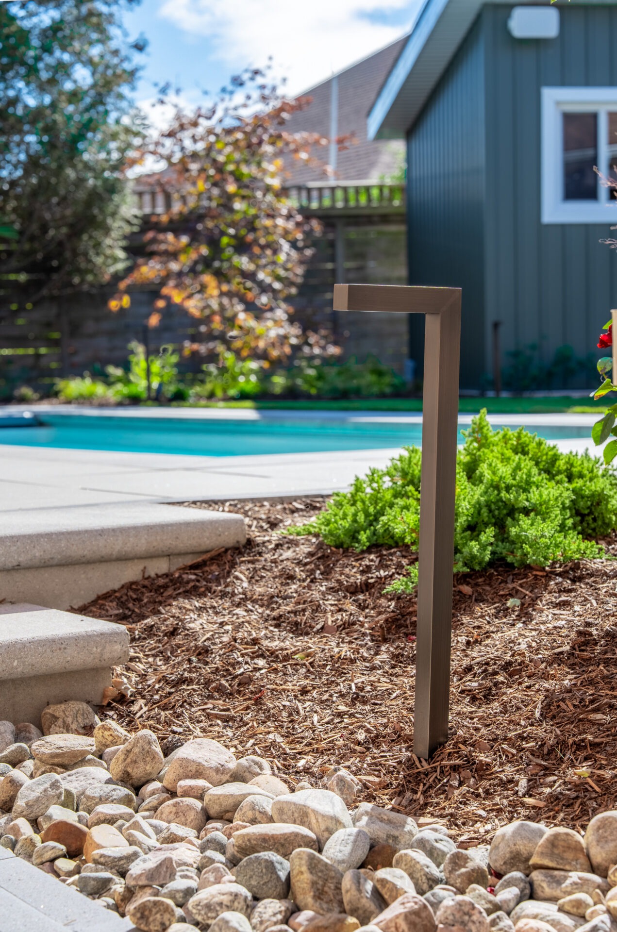 A low-angle shot of a modern, bronze-colored L-shaped path light installed in a mulched garden bed. In the background, a swimming pool and a dark green outbuilding are visible under a bright, partly cloudy sky.