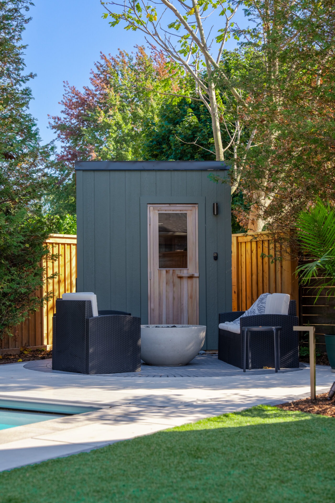Modern green backyard sauna with wood door next to a round fire pit and wicker lounge chairs.