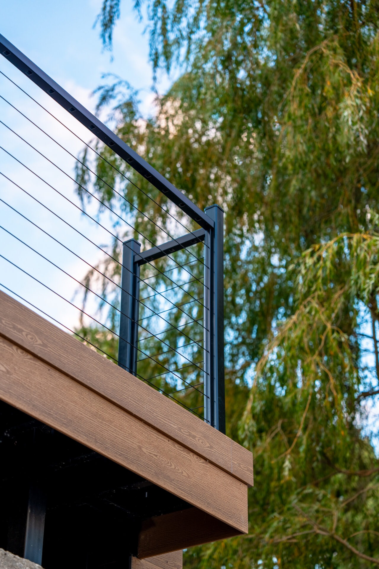 Upward view of a deck's wood-textured fascia and cable railing system set against green trees.
