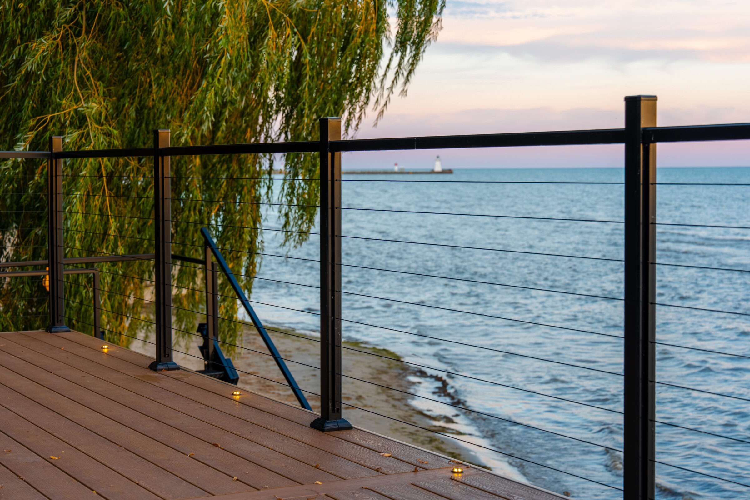 Deck view through cable railings toward a distant lighthouse on the water during sunset.