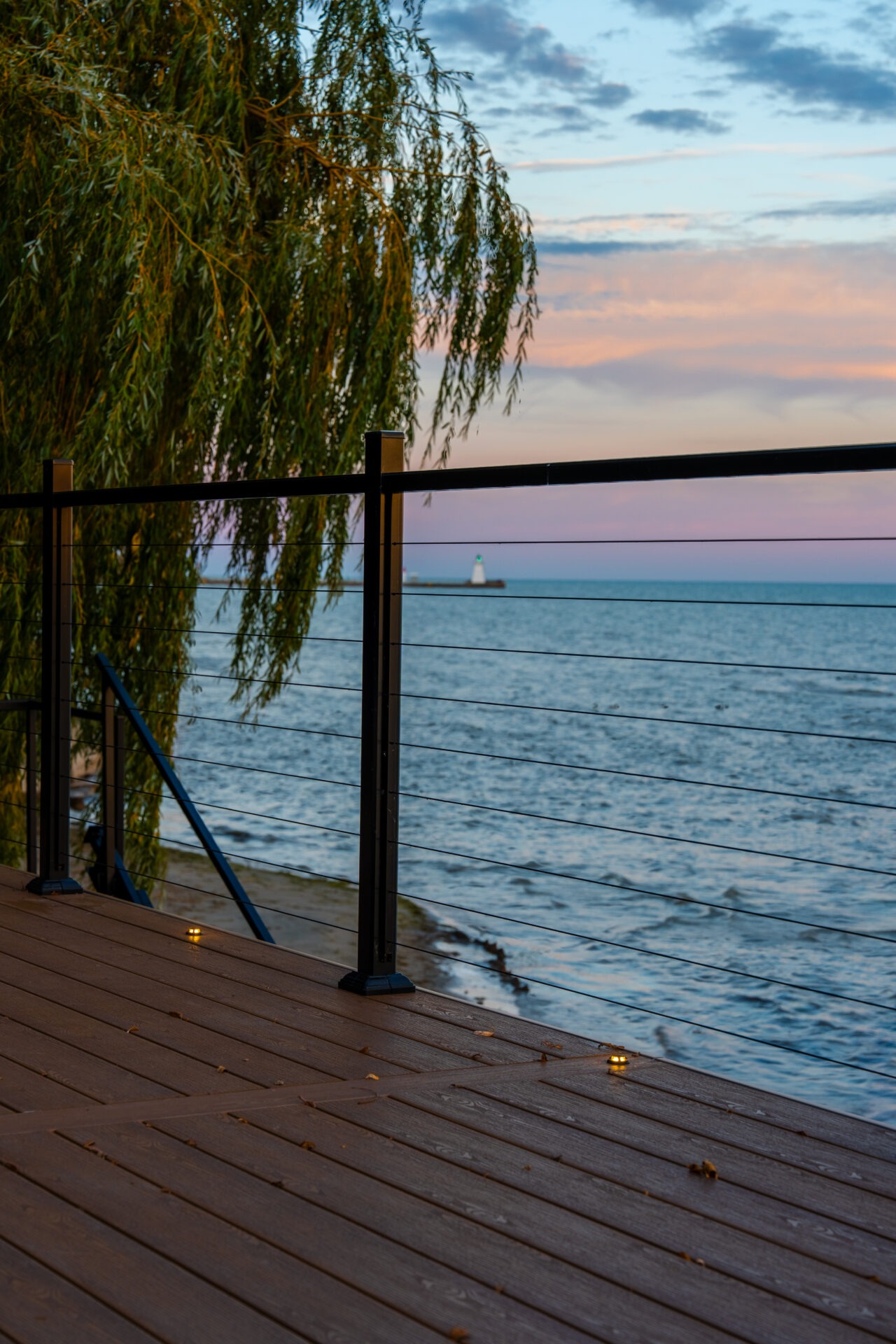 Modern deck with cable railings and built-in floor lighting overlooking a calm lake at dusk.