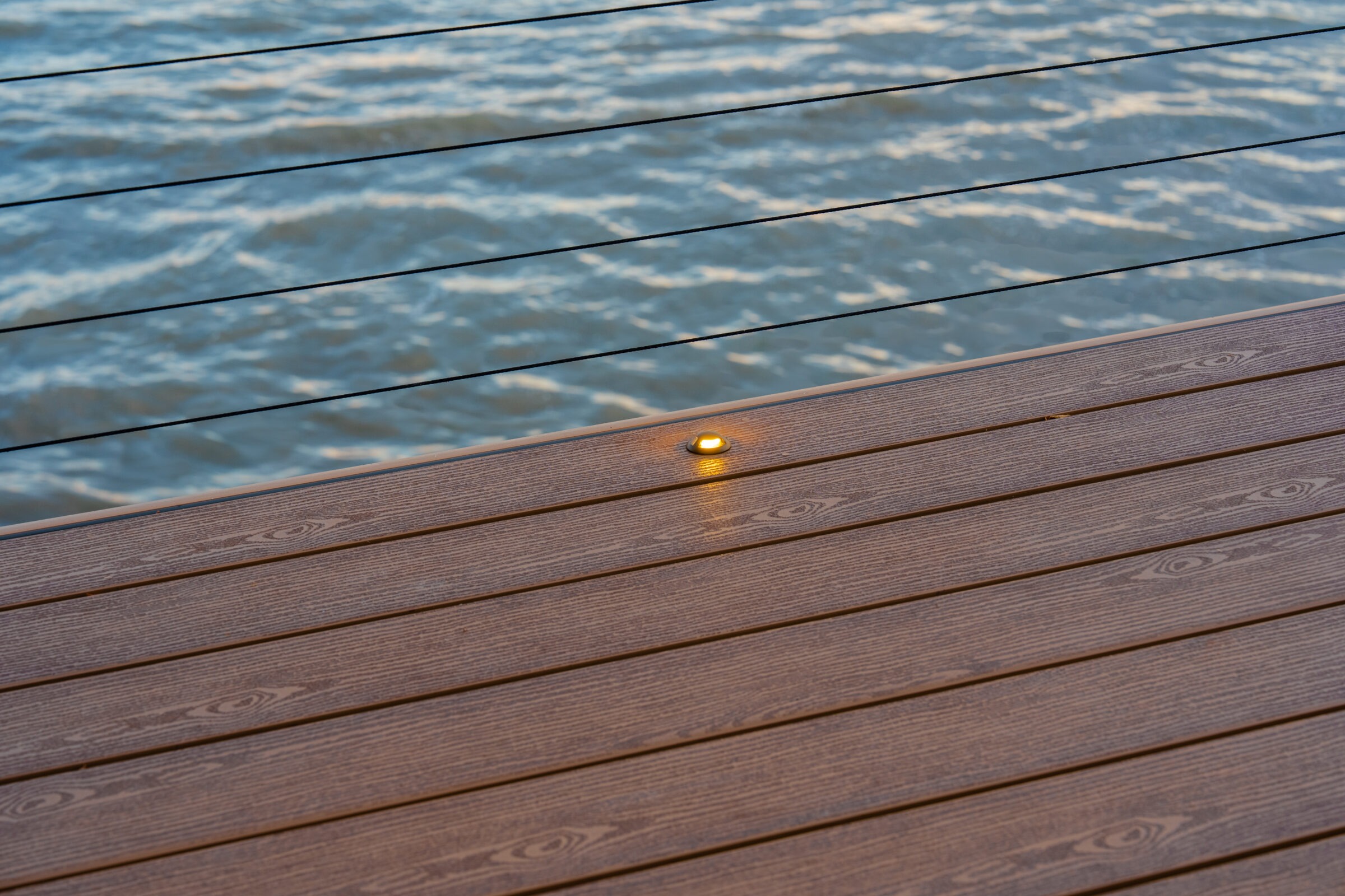 Close-up of a recessed LED light in a wood-plank deck overlooking blue water.