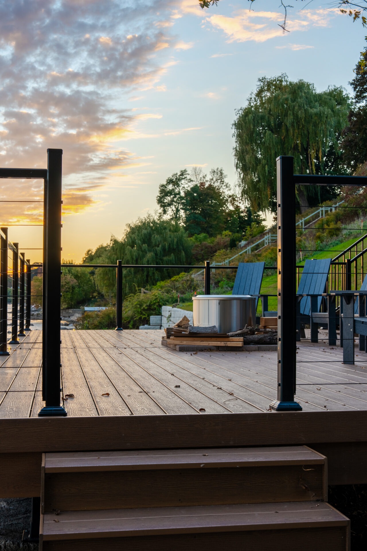 A wood-textured deck with black cable railings and a fire pit area during a golden sunset.