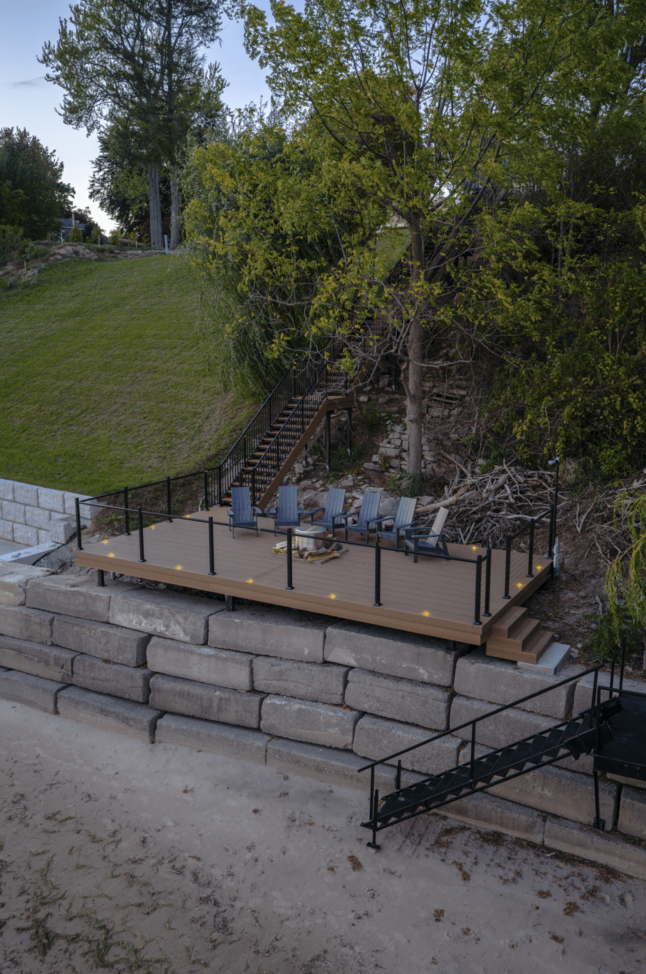 Side view of a shoreline deck built on a stone block wall, featuring seating, a fire pit, and stairs.