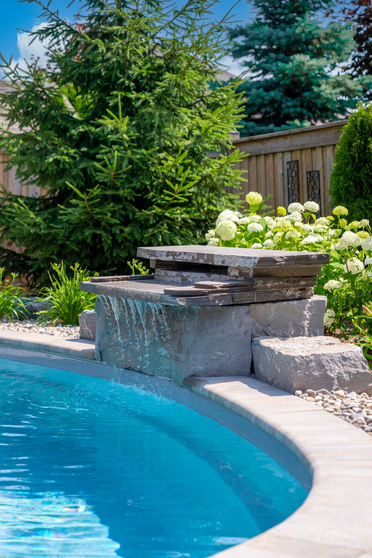 Close-up of a custom natural stone waterfall feature spilling into a clear blue swimming pool, featuring smooth flagstone surfaces and surrounded by white hydrangeas and decorative river rock.