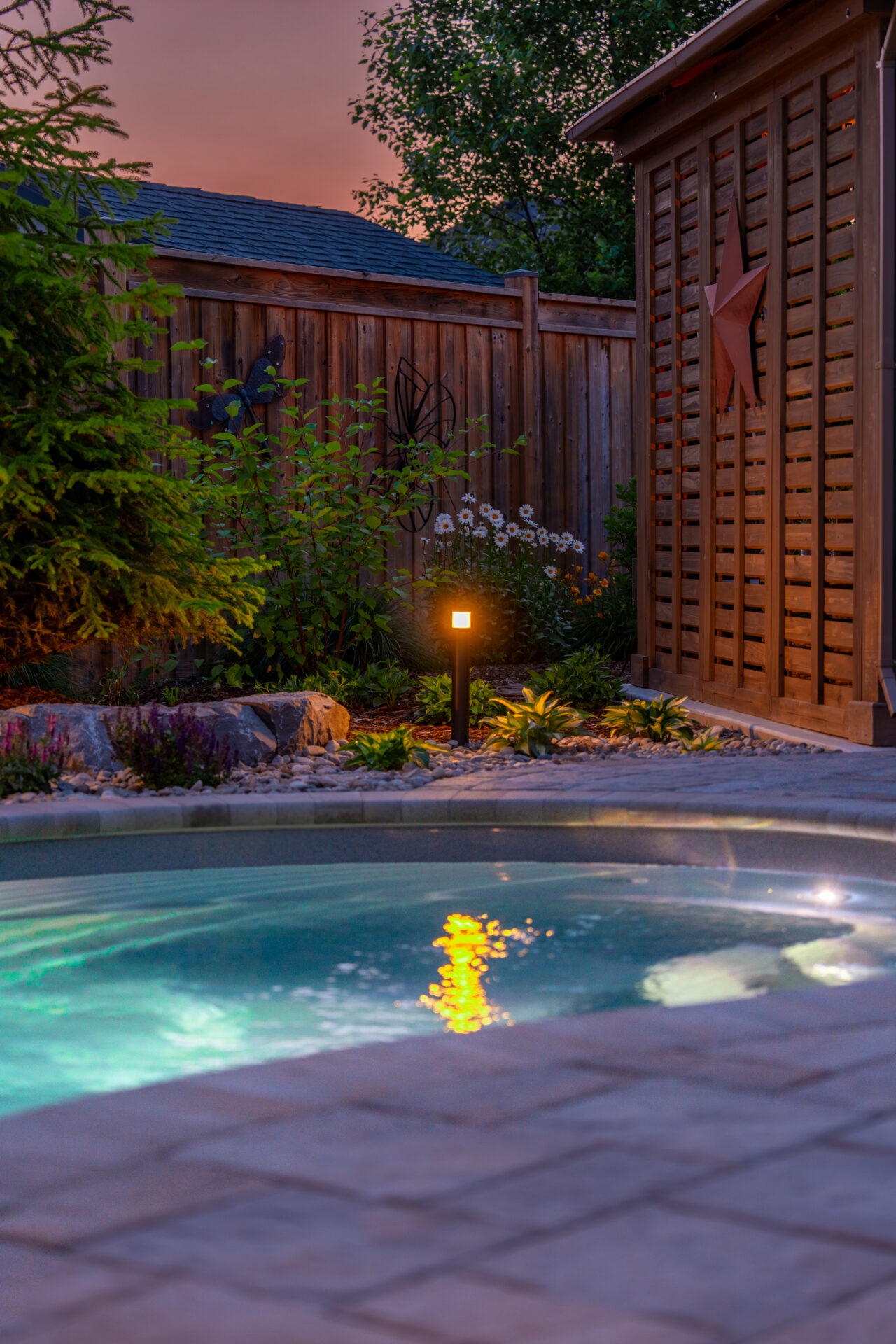 Twilight view of a glowing custom pool in Port Dover with integrated landscape lighting and a privacy screen, highlighting durable backyard heating options.