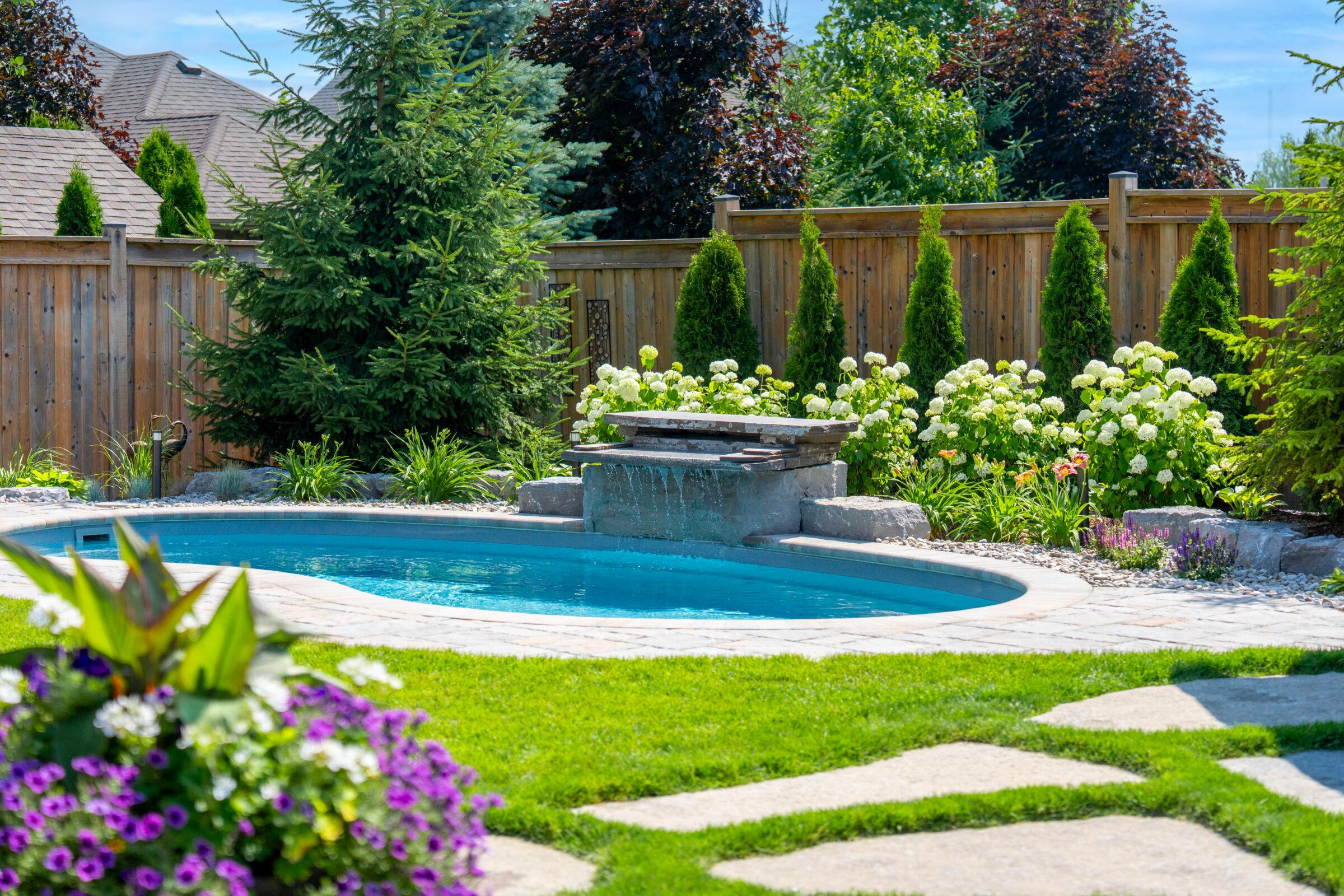 A luxury curved inground swimming pool by Doverscapes featuring a natural stone waterfall feature, surrounded by white hydrangeas, lush green lawn, and a privacy fence.