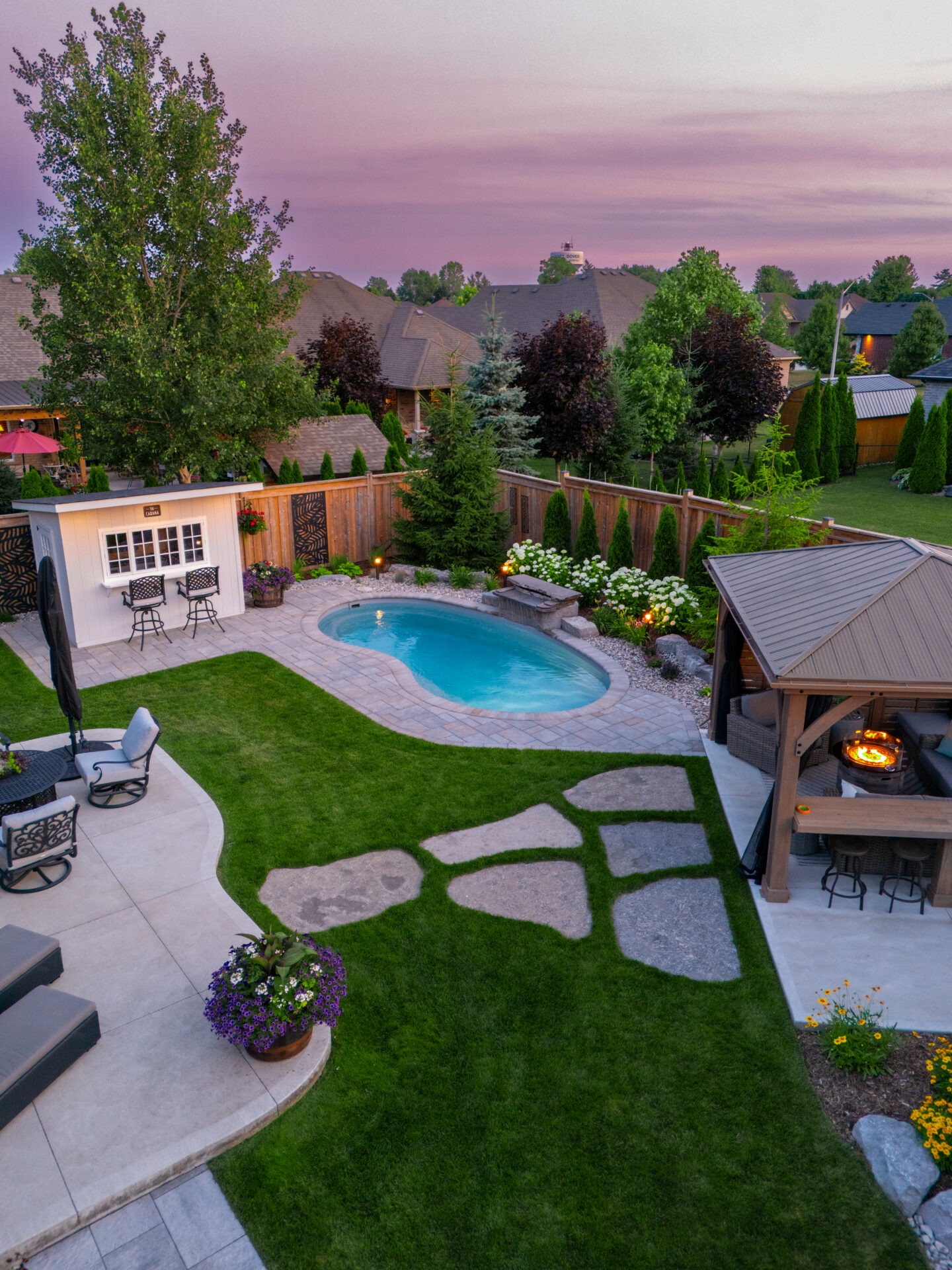 Aerial sunset view of a custom curved inground pool with a stone waterfall, a poolside cabana bar, and a covered lounge area with a fire pit, designed and installed by Doverscapes in Port Dover.