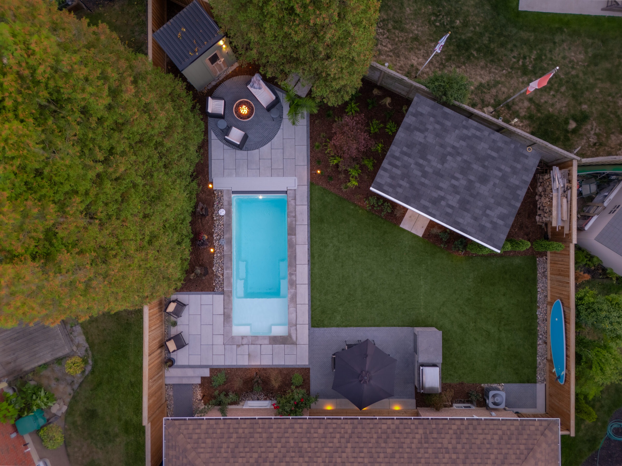 Aerial view of a modern luxury backyard featuring a rectangular plunge pool, a circular stone fire pit area with seating, and a manicured green lawn.