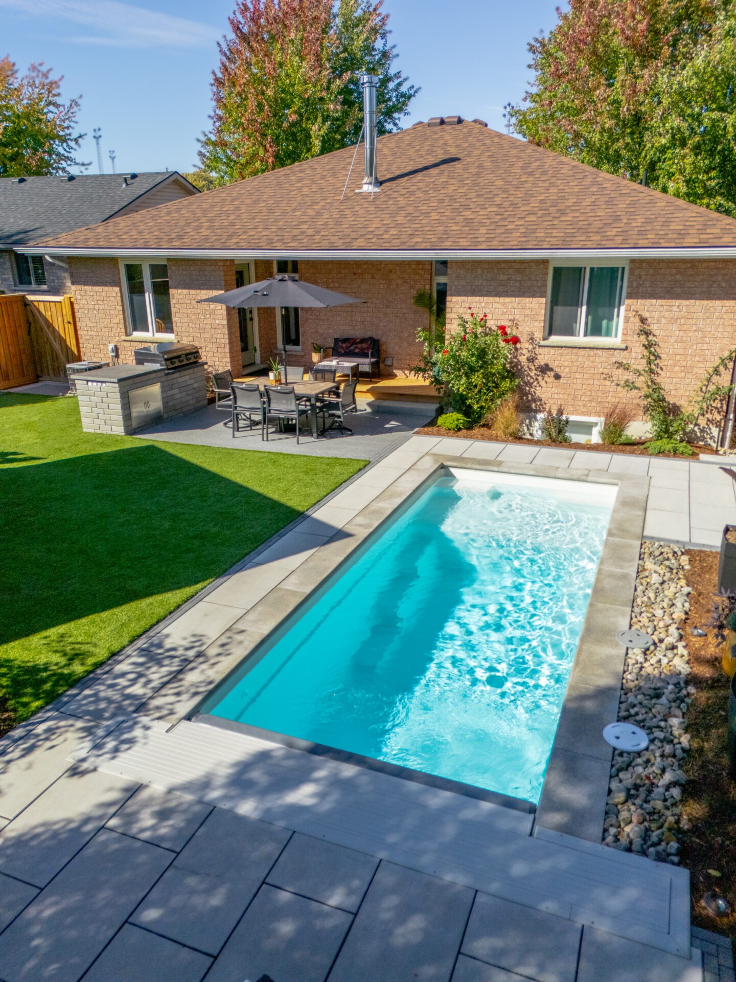 Backyard view of a brick bungalow featuring a modern outdoor kitchen with a built-in grill, a stone patio dining area with a black umbrella, and a rectangular white-lined plunge pool surrounded by grey pavers.