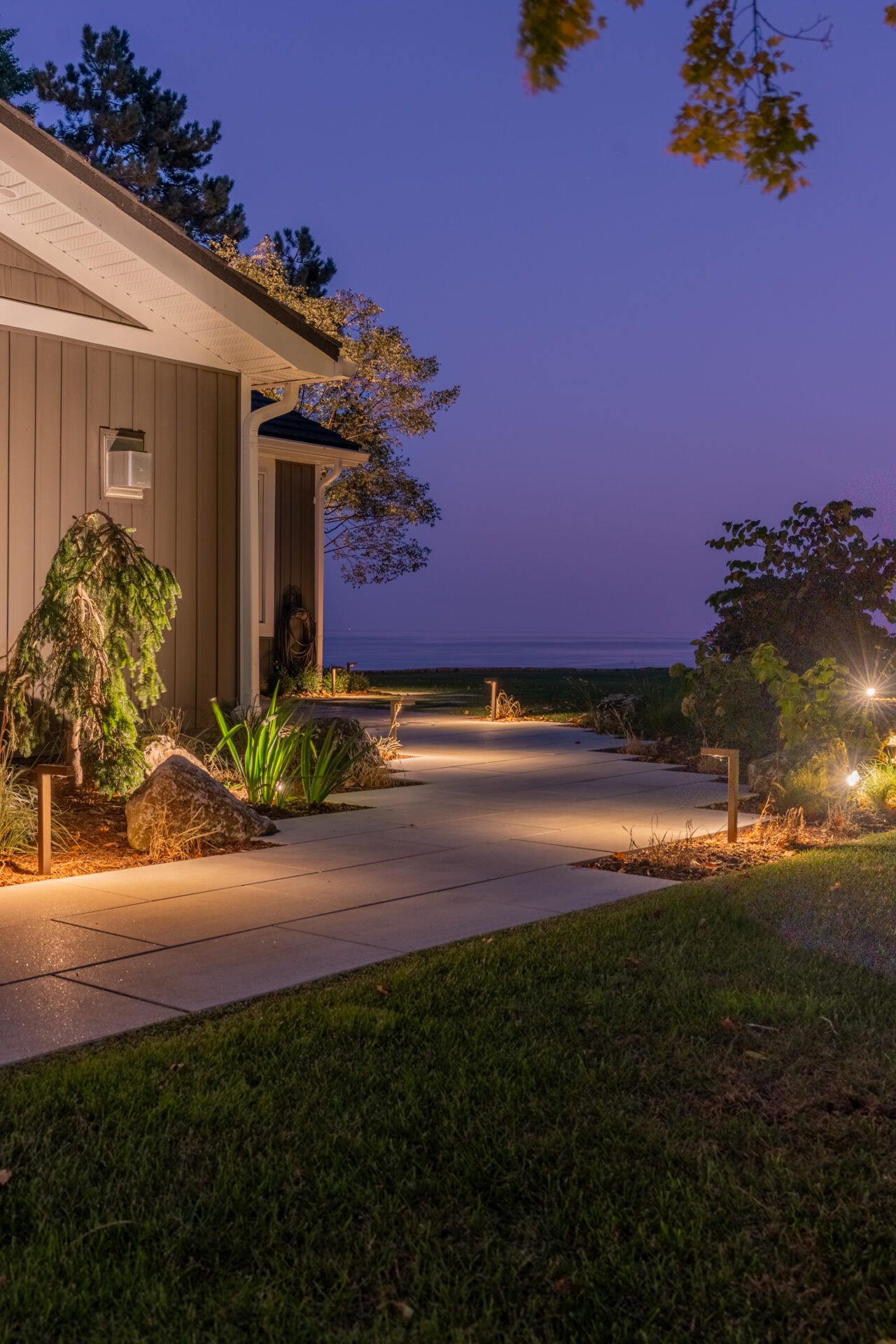 A modern stone walkway at twilight illuminated by strategic low-voltage landscape path lights, leading toward a waterfront property with subtle architectural lighting on the house exterior.